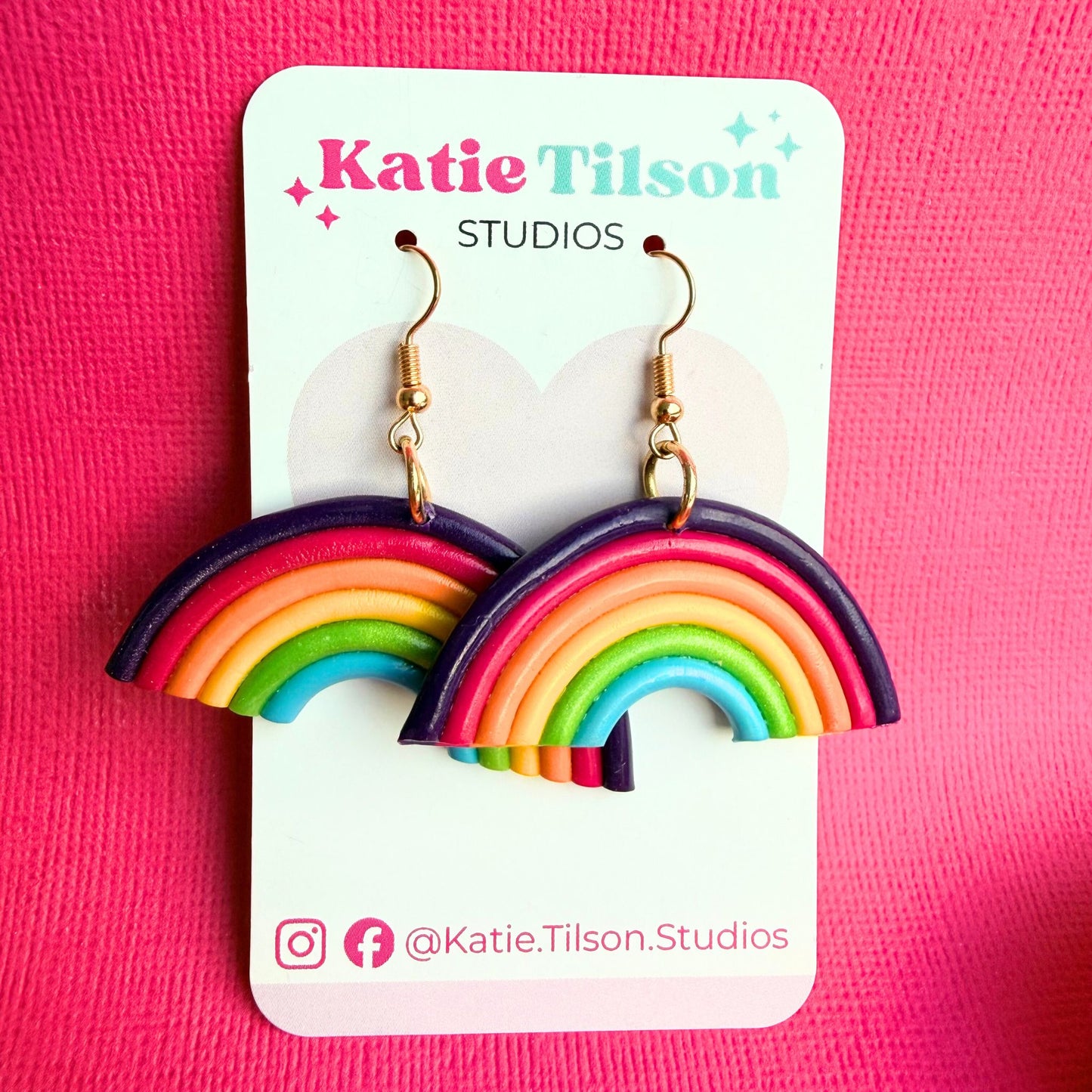Rainbow-shaped dangle earrings on a Katie Tilson Studios card against a pink background.