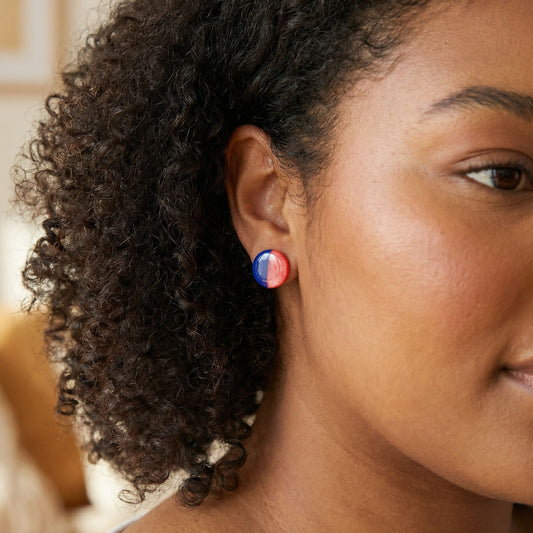 Close-up of a woman wearing round cobalt blue and coral half-and-half circular stud earrings with a glossy finish against a blurred background.