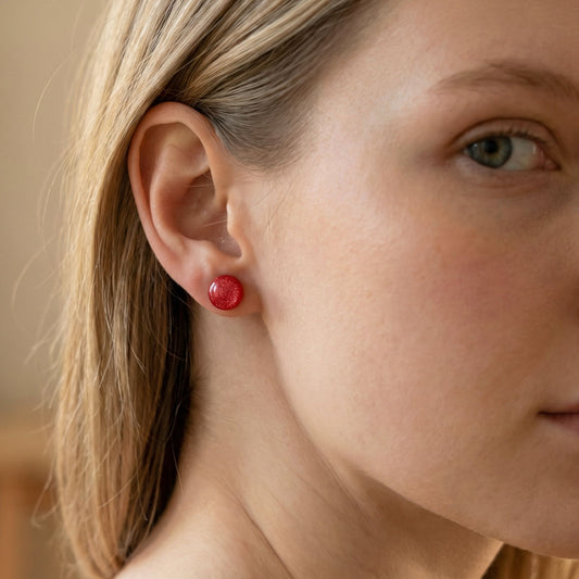 Close-up of a woman wearing round coral stud earrings with a glossy finish against a blurred background.