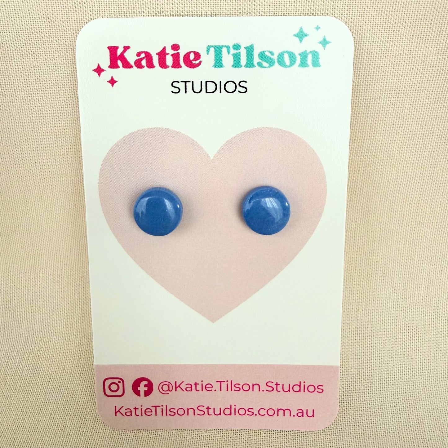 Cornflower blue circular stud earrings on a Katie Tilson Studios card against a beige background.
