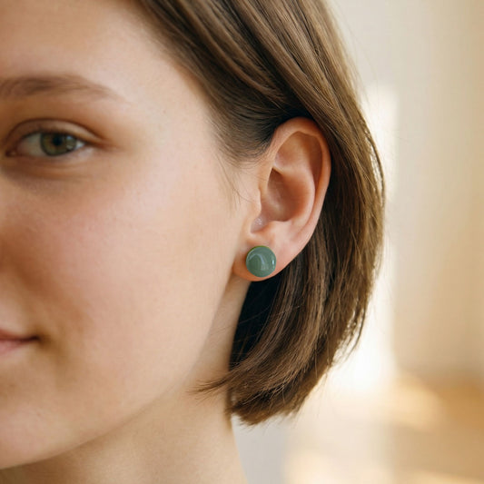 Close-up of a woman wearing round eucalyptus green stud earrings with a glossy finish against a blurred background.