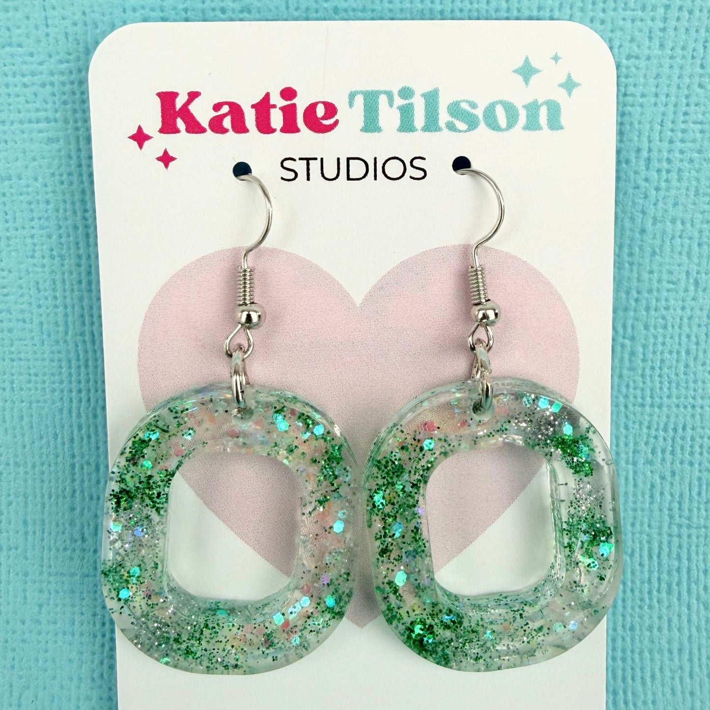 Green glittery hoop shaped dangle earrings on a Katie Tilson Studios card against a blue background.