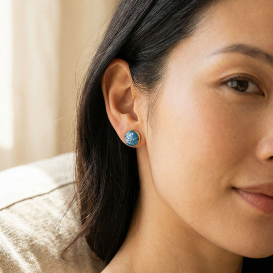 Close-up of a woman wearing teal stud earrings with gold details.