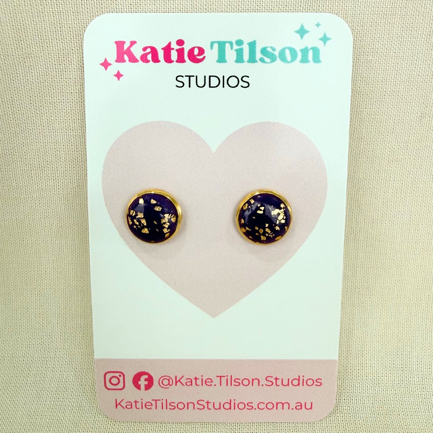 Purple handmade stud earrings with gold detail on a Katie Tilson Studios packaging with a beige background.