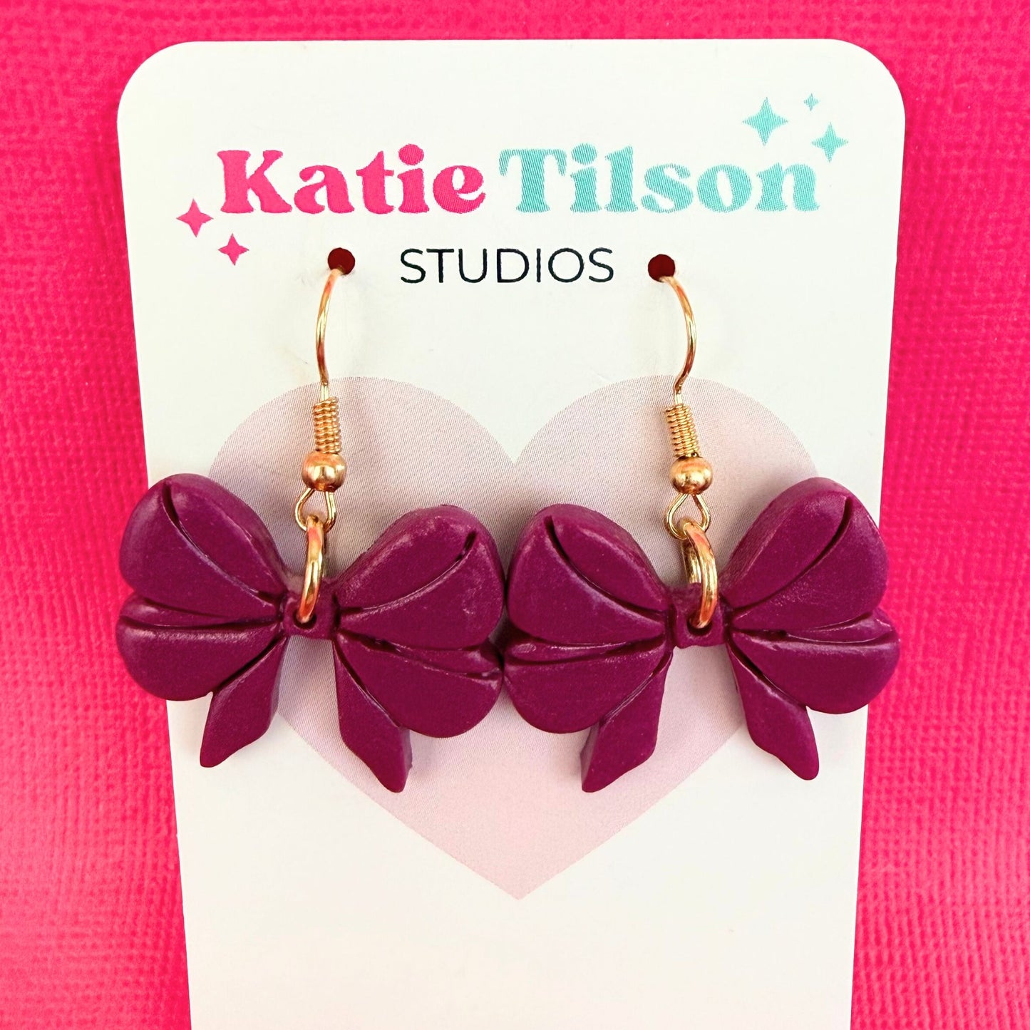 Purple bow-shaped dangle earrings on a Katie Tilson Studios card against a pink background.