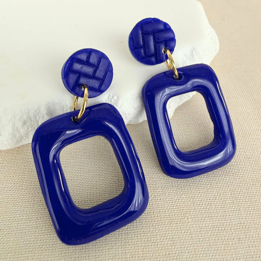 Cobalt blue frame drop earrings with a glossy finish hanging from matte blue textured studs against an oat-coloured background.