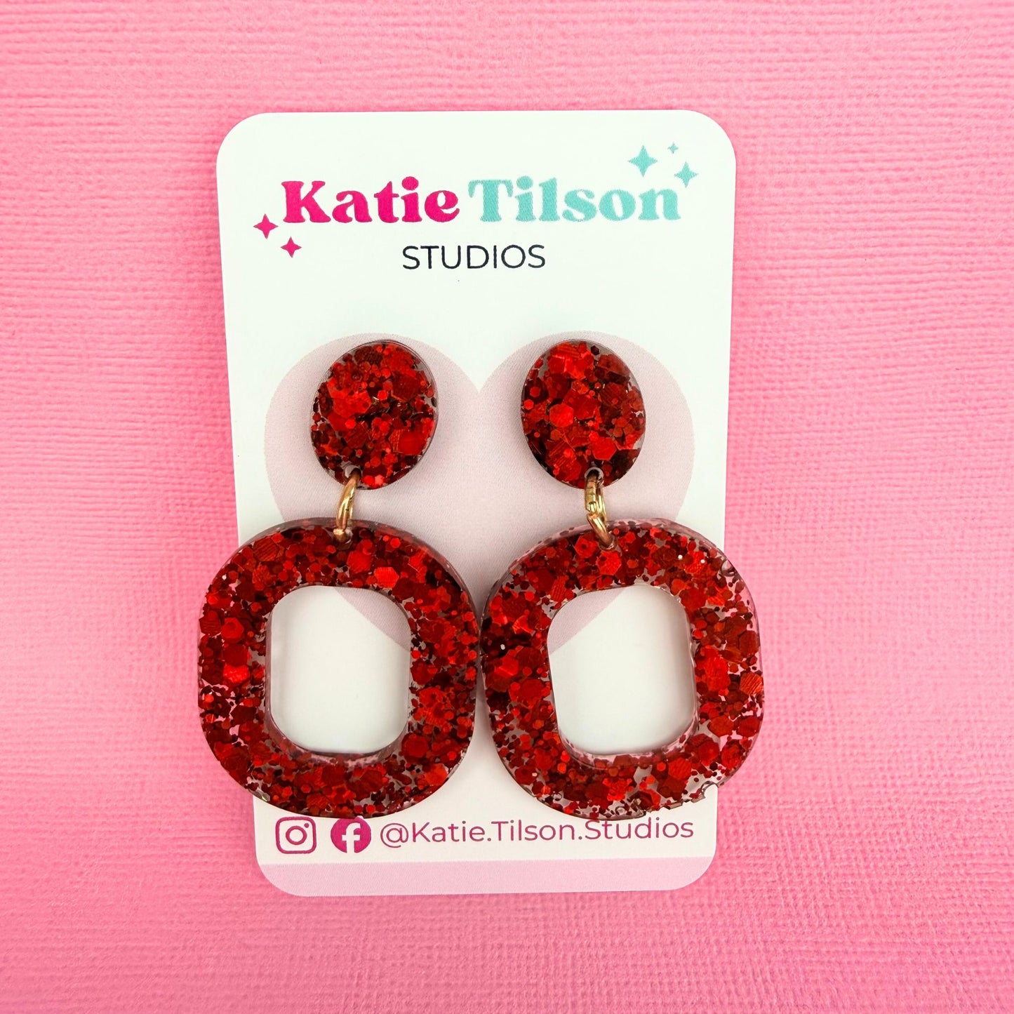 Ruby Glow red glittery resin drop earrings on a Katie Tilson Studios card against a pink background.