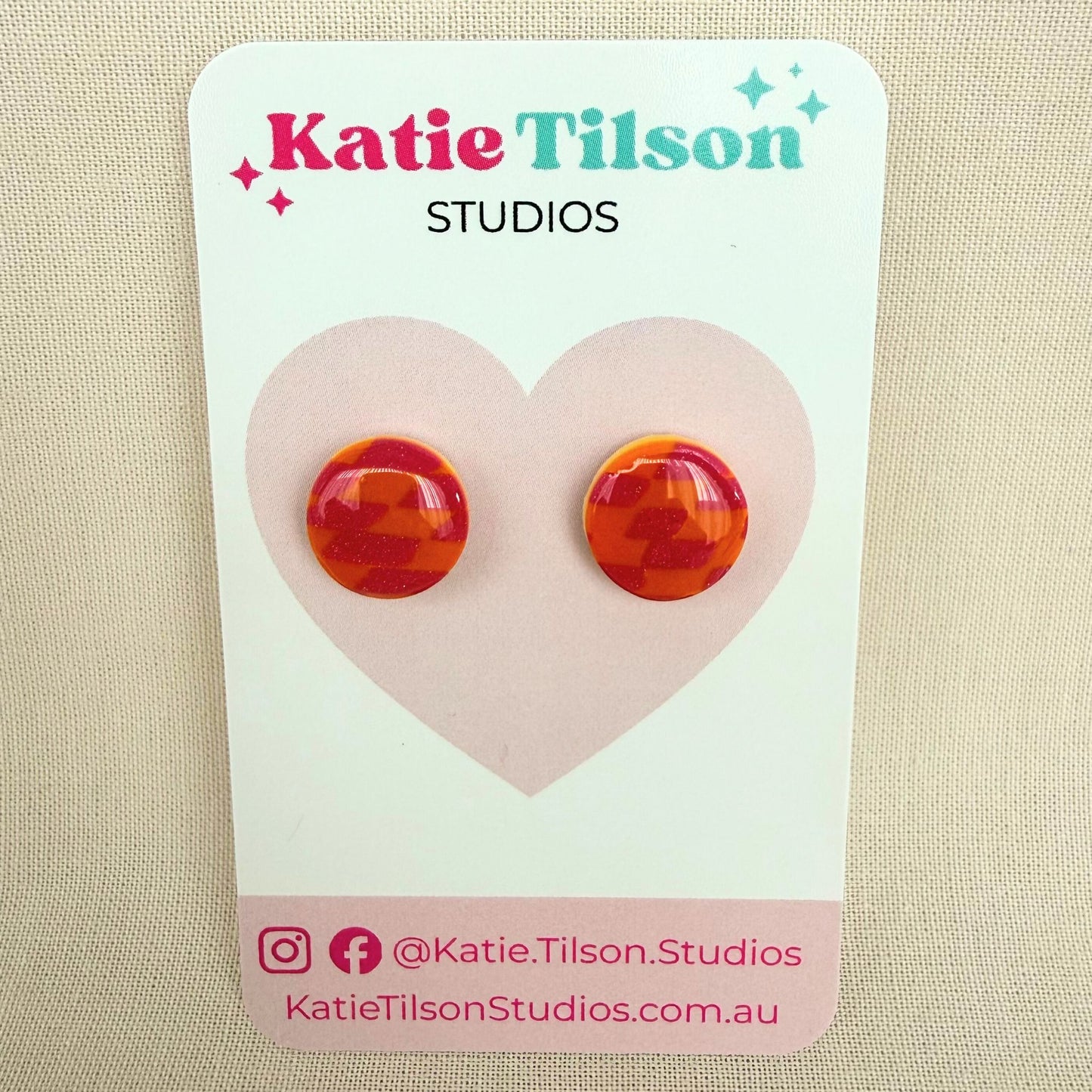 Pink and orange check circular stud earrings on a branded earring card against a beige background.