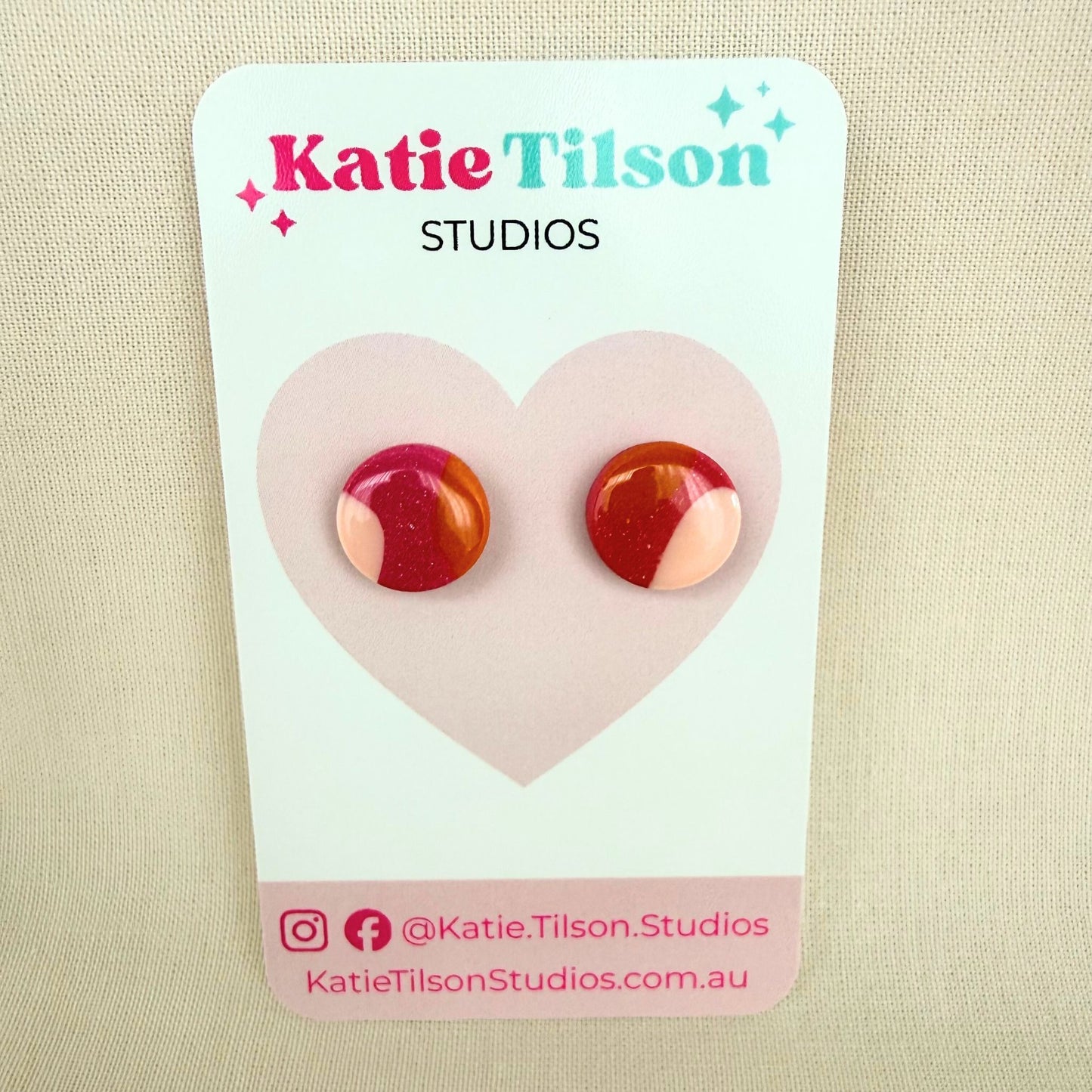 Pair of dark pink, orange and pastel pink colorful stud earrings on a Katie Tilson Studios card against a beige background.