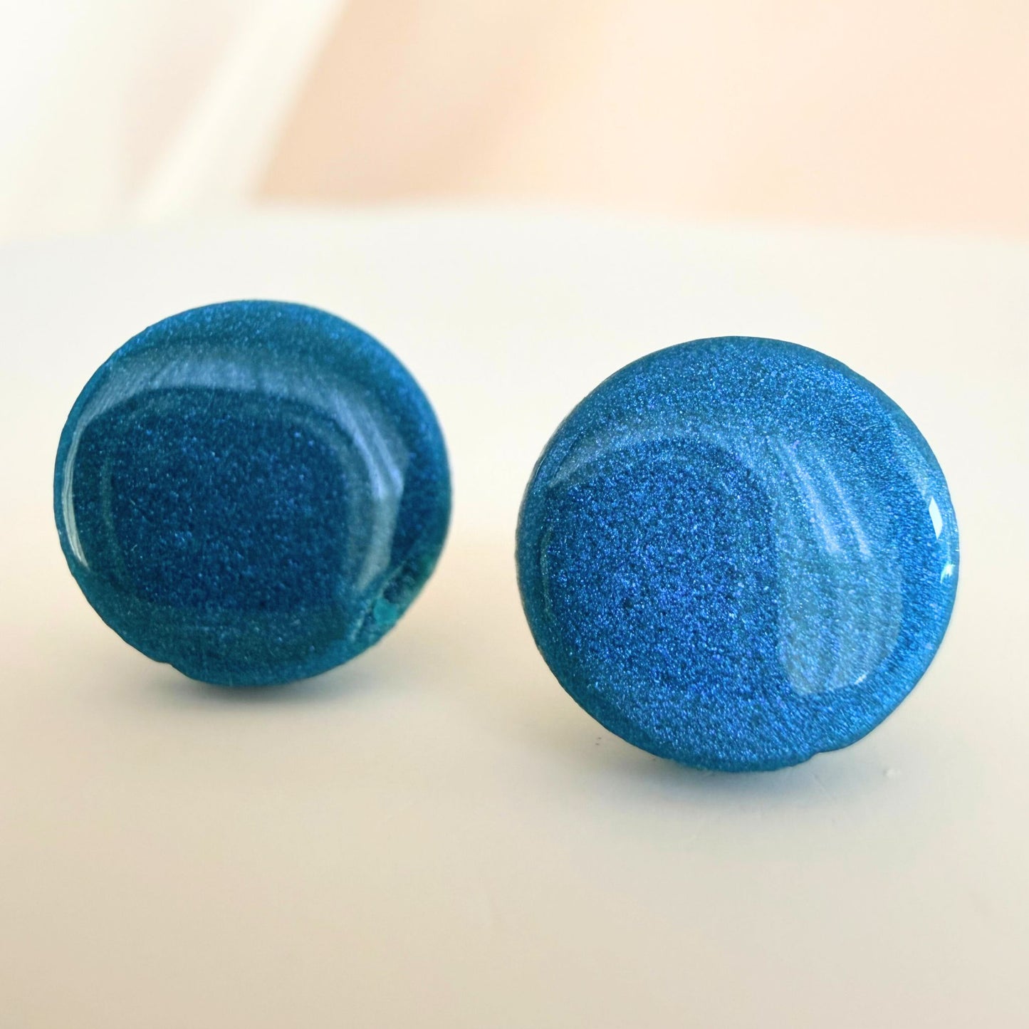 A pair of teal circle studs with shimmer and glossy resin finish resting on a white surface.