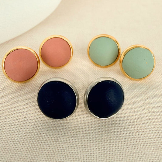 Round stud earrings in navy, blush, and soft green, set in gold and silver-tone bezels, arranged on neutral fabric.