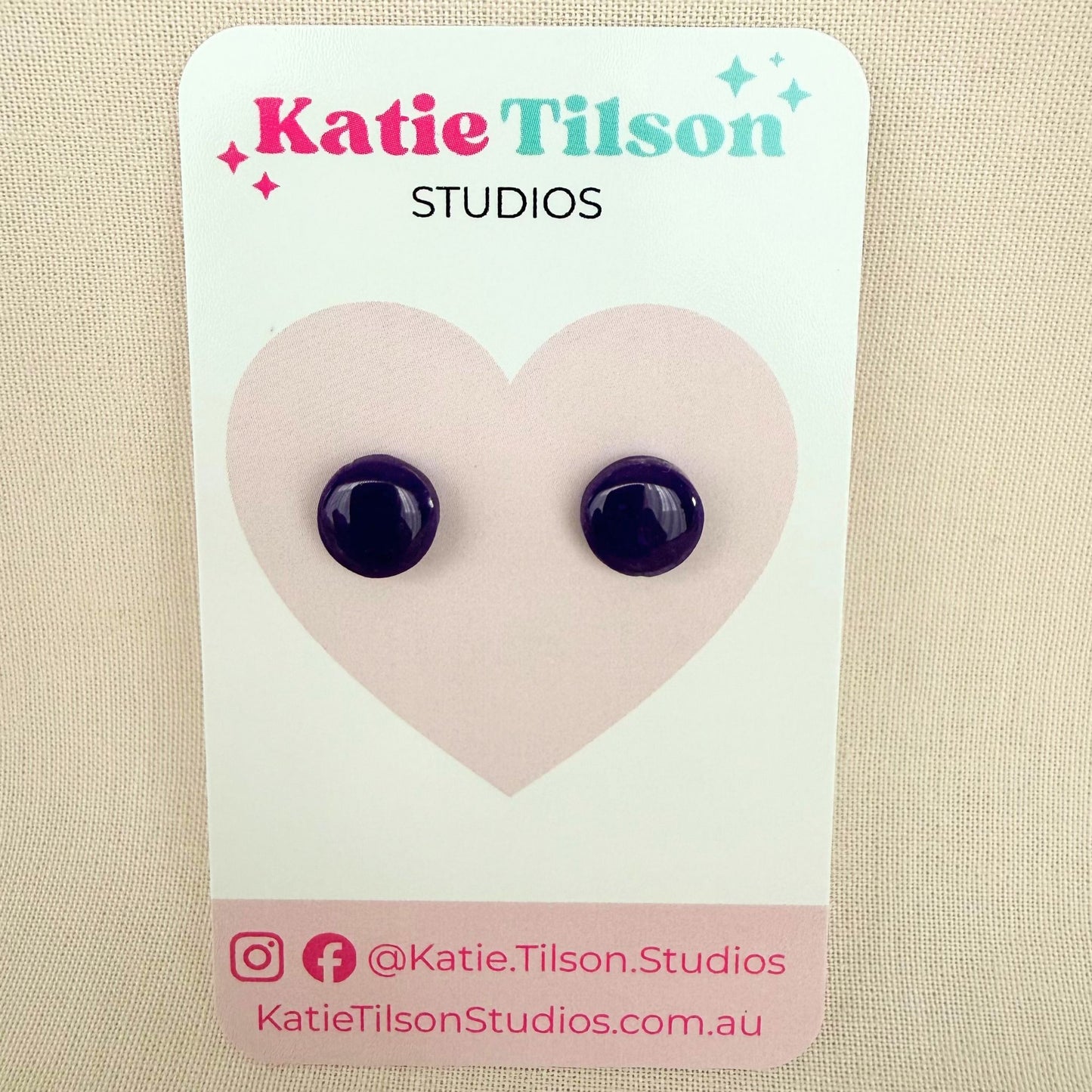 Deep violet purple circular studs on a branded earring card against a beige background.