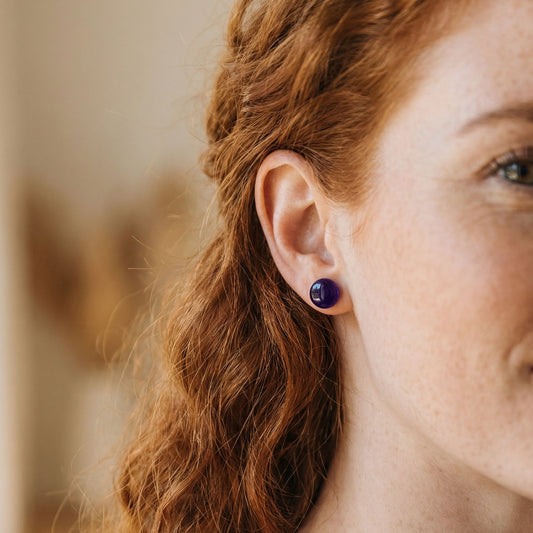 Close-up of a woman wearing violet purple stud earrings with a glossy finish against a blurred background.