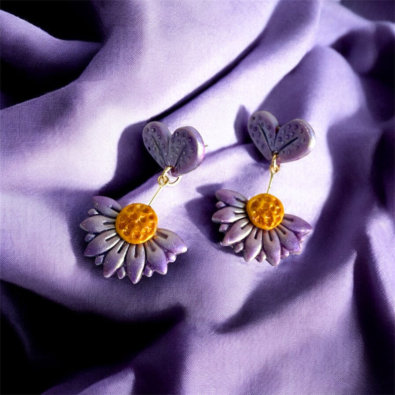 a pair of handmade daisy shaped polymer clay stud earrings in purple with yellow centres on a purple fabric background