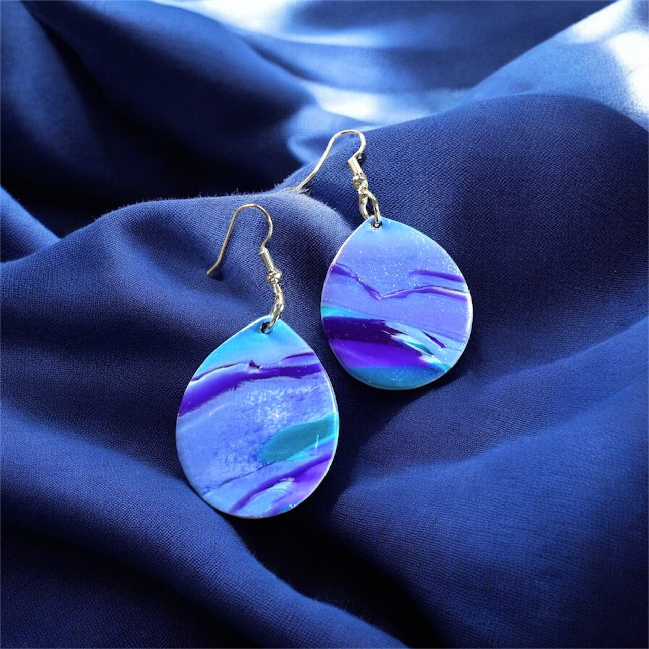 a pair of blue and purple marbled dangle polymer clay earrings in a teardrop shape on a blue fabric background