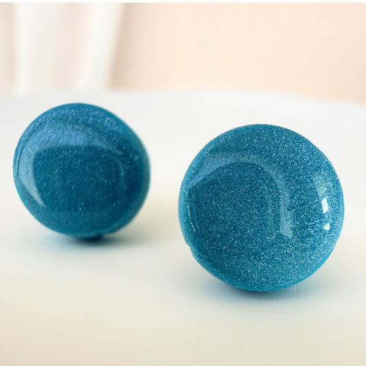 A pair of bright aqua circle studs with shimmer and glossy resin finish resting on a white surface.