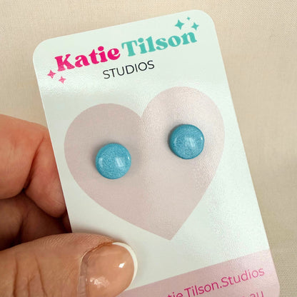Aqua blue circular stud earrings on a Katie Tilson Studios card held by a hand.