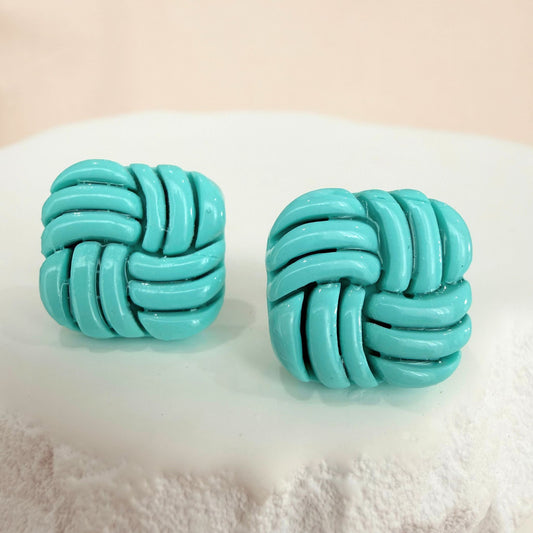 Aqua knotted stud earrings on a textured white surface with a light beige background.