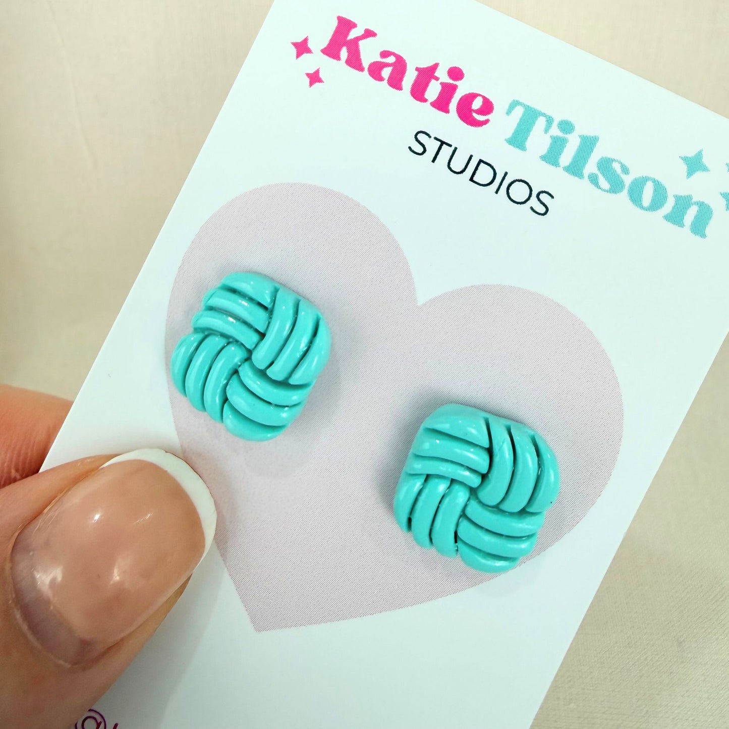 Aqua knotted stud earrings on a Katie Tilson Studios card held by a hand.