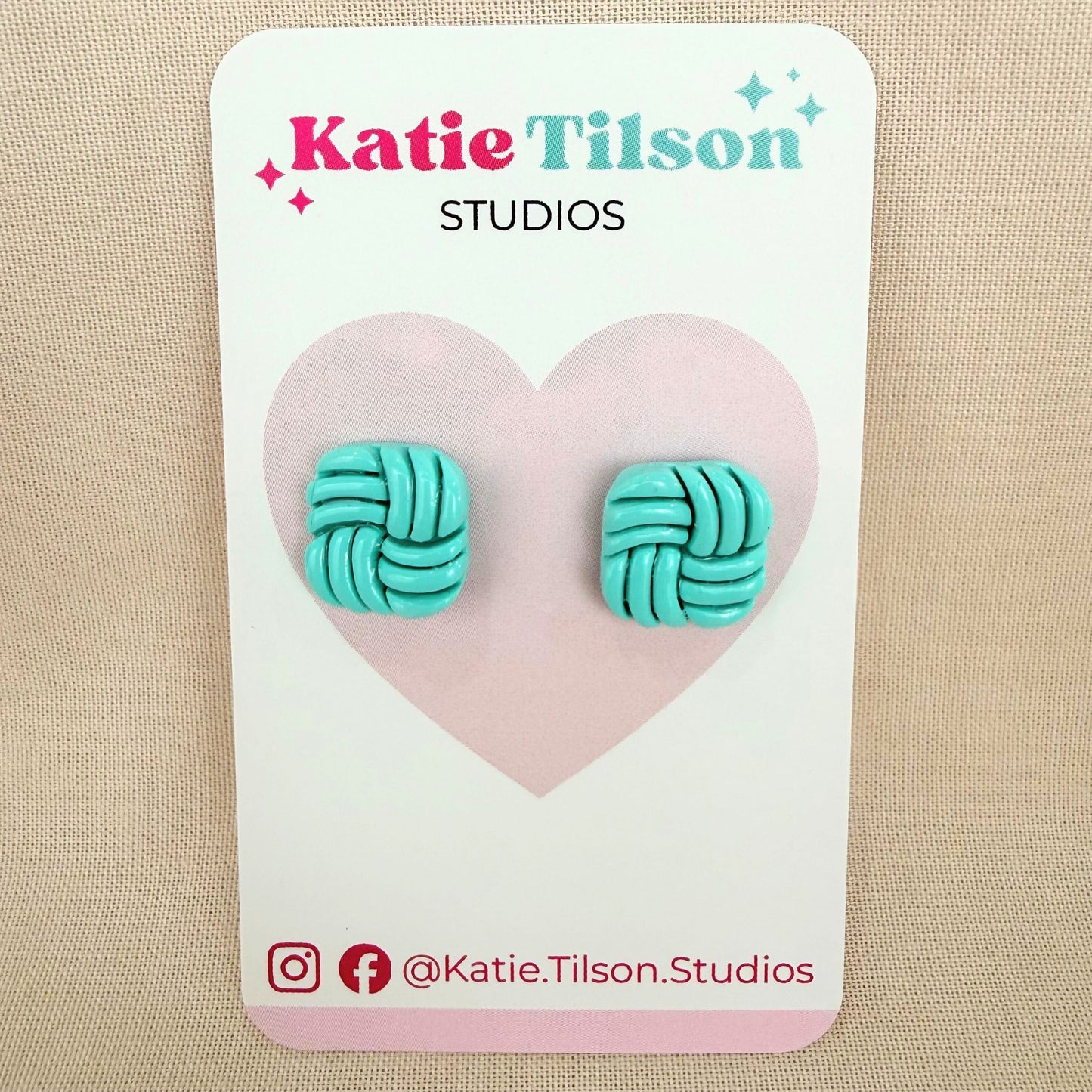 Aqua knotted stud earrings on a textured white surface with a light beige background.