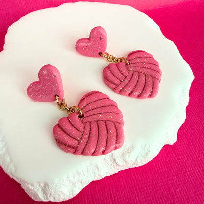 Pink heart-shaped drop earrings with knitted texture and subtle shimmer on a white stone with a pink background.