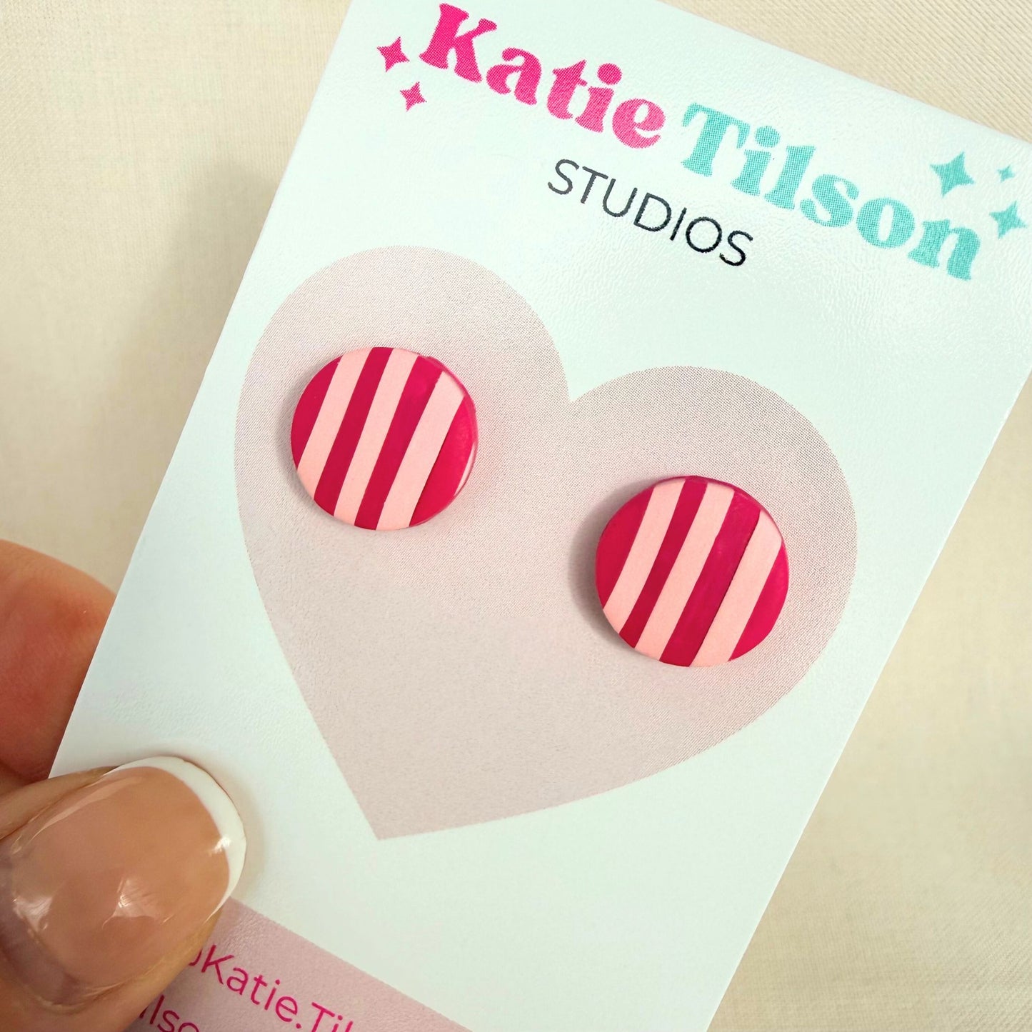 A pair of pink striped circular stud earrings on a Katie Tilson Studios branded card held by a hand.