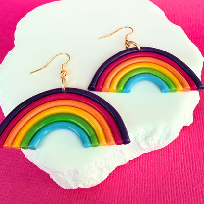Australian handmade rainbow-shaped earrings in bright rainbow colours with gold hooks on a white stone with a pink background.