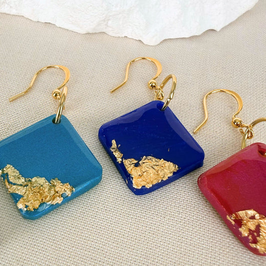 Three pairs of square earrings in cobalt, magenta and teal with gold accents and high shine finish on a textured surface.