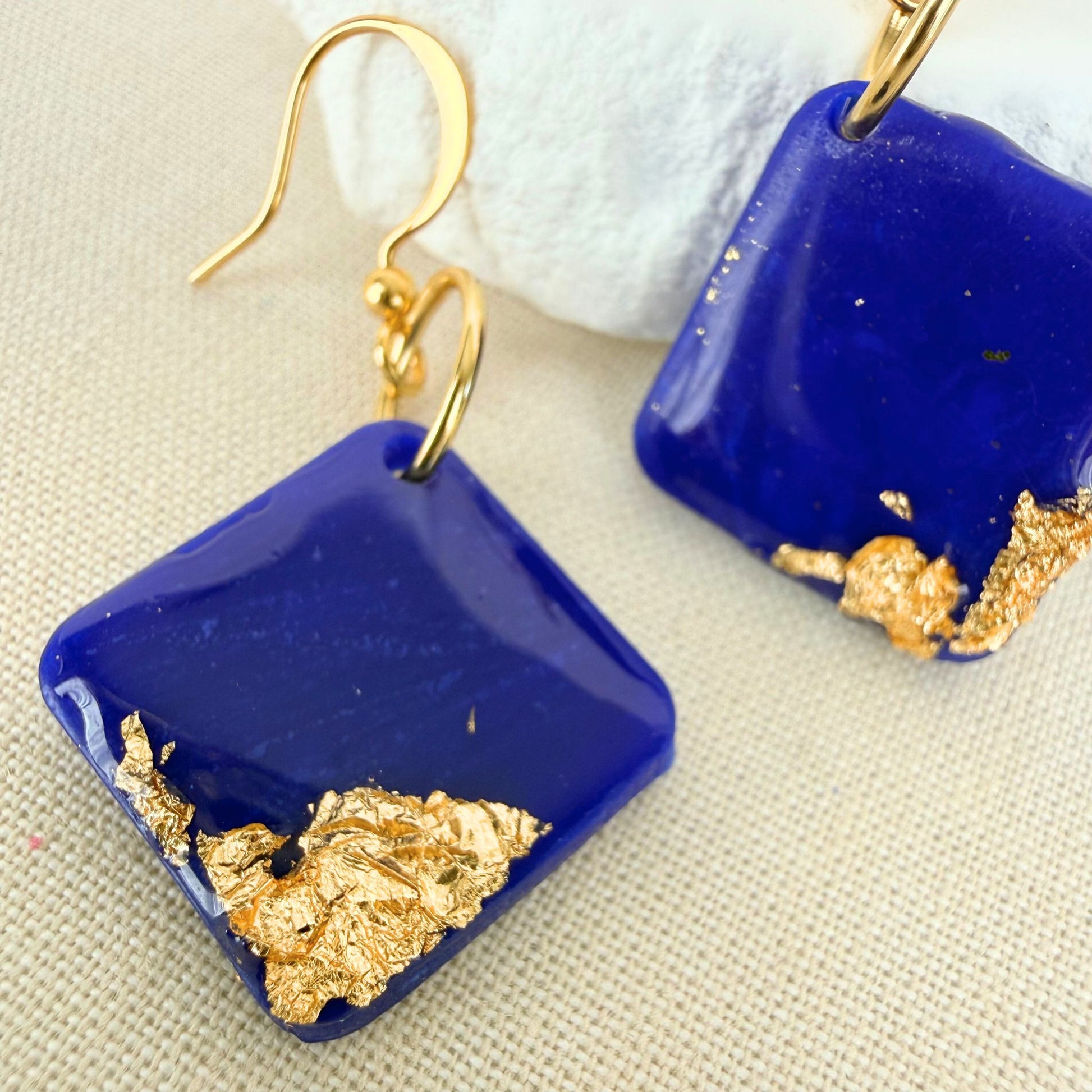 Close up of cobalt blue square earrings with gold accents on a beige fabric background.