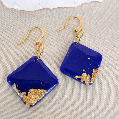 Cobalt blue square earrings with gold leaf accents and high gloss shine on a beige fabric background.