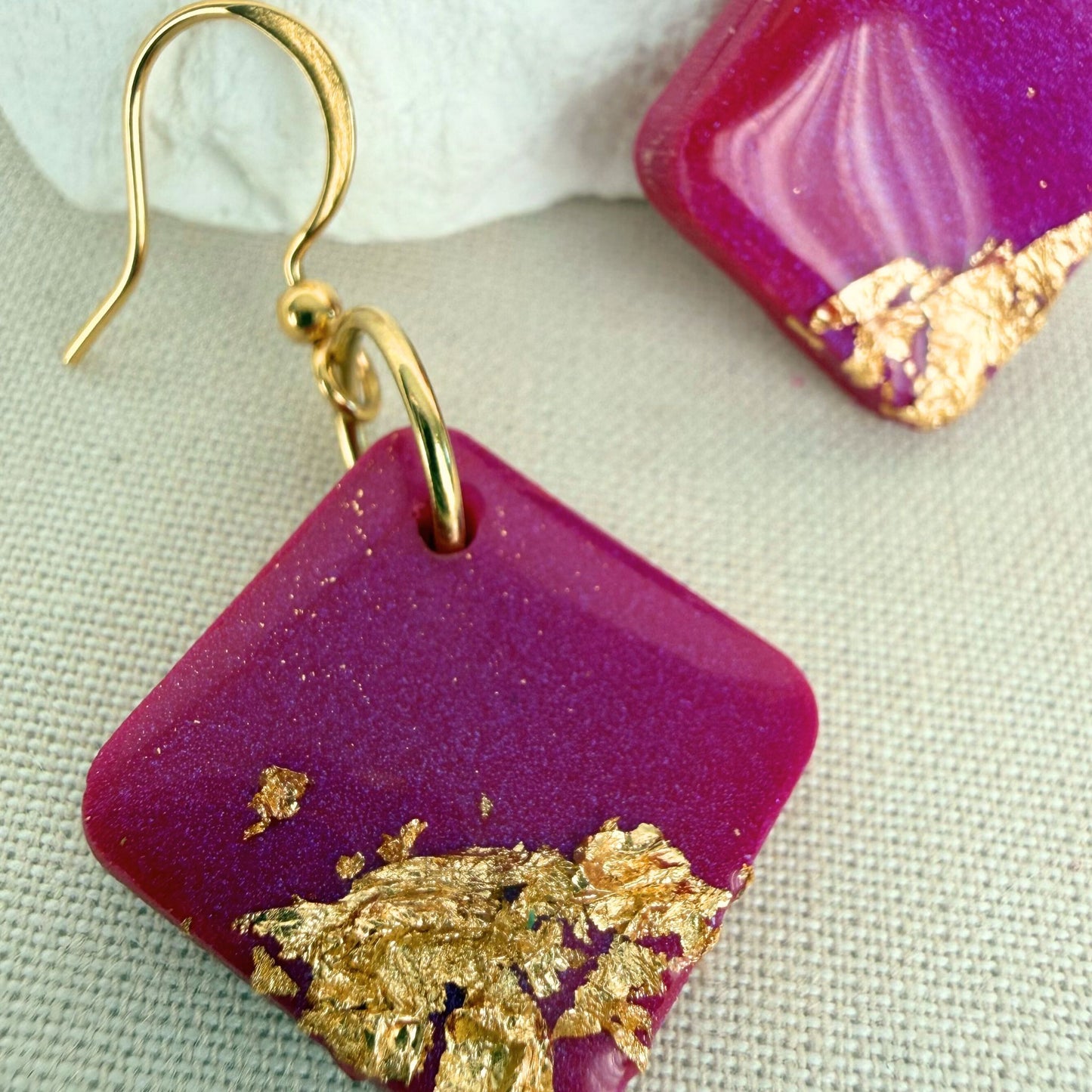 Close up of magenta dangle earrings with gold leaf on a light beige fabric background.