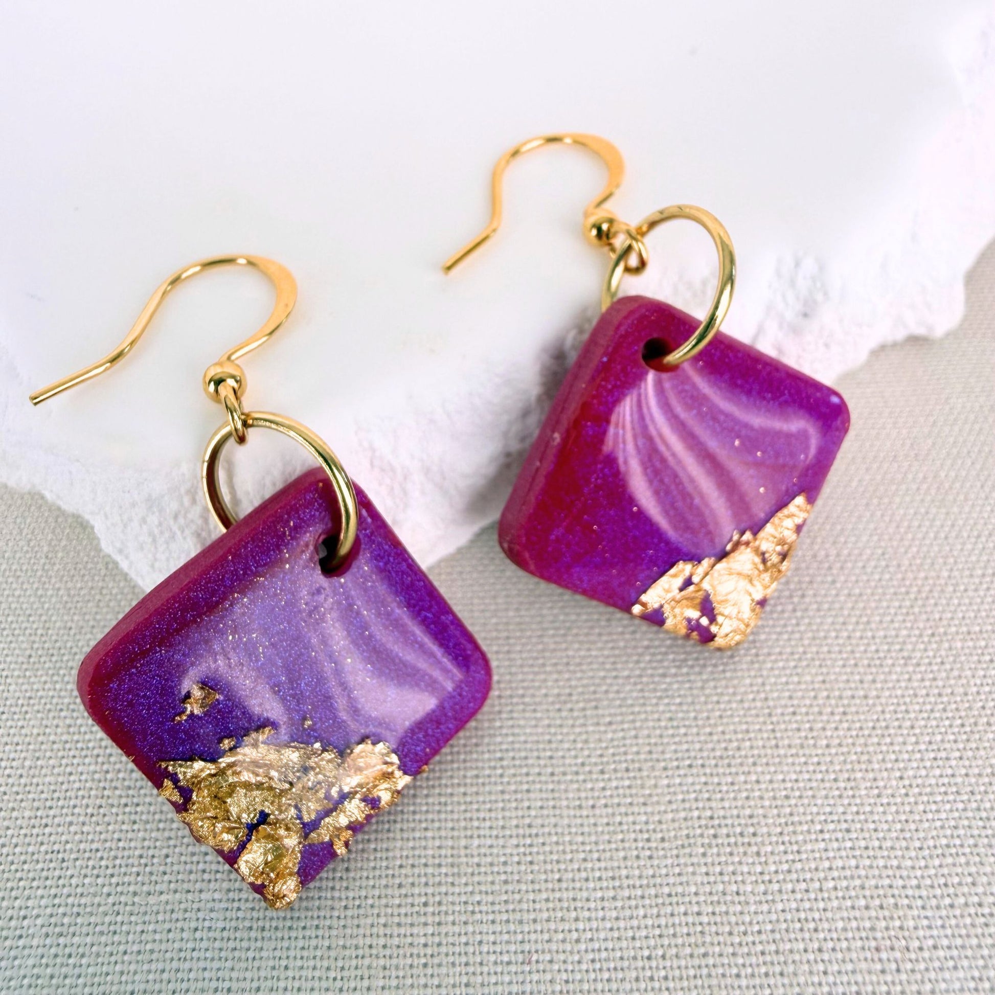 Magenta colour square dangle earrings with gold flakes on a textured surface.
