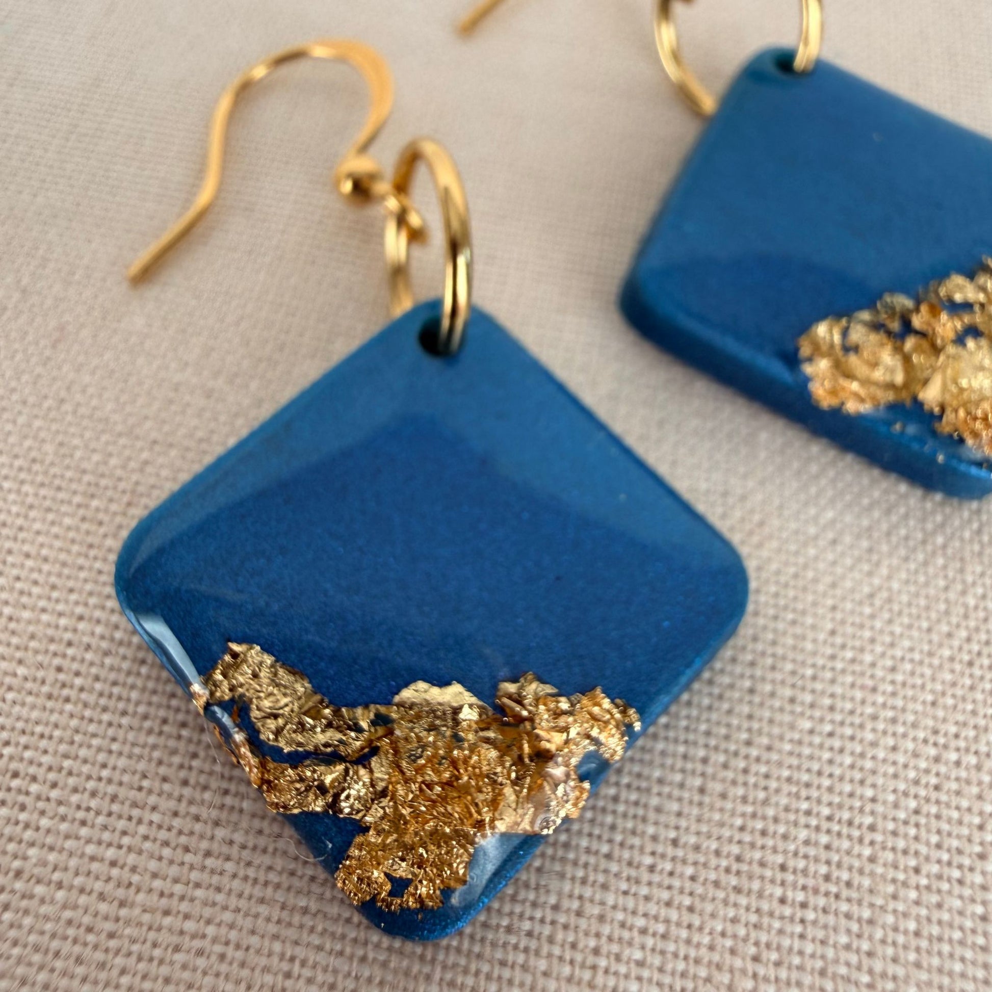 Teal square dangle earrings in high gloss with gold leaf on a beige fabric background.
