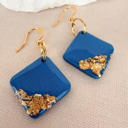 Teal blue square handmade earrings with gold leaf and glossy finish on a beige fabric background.