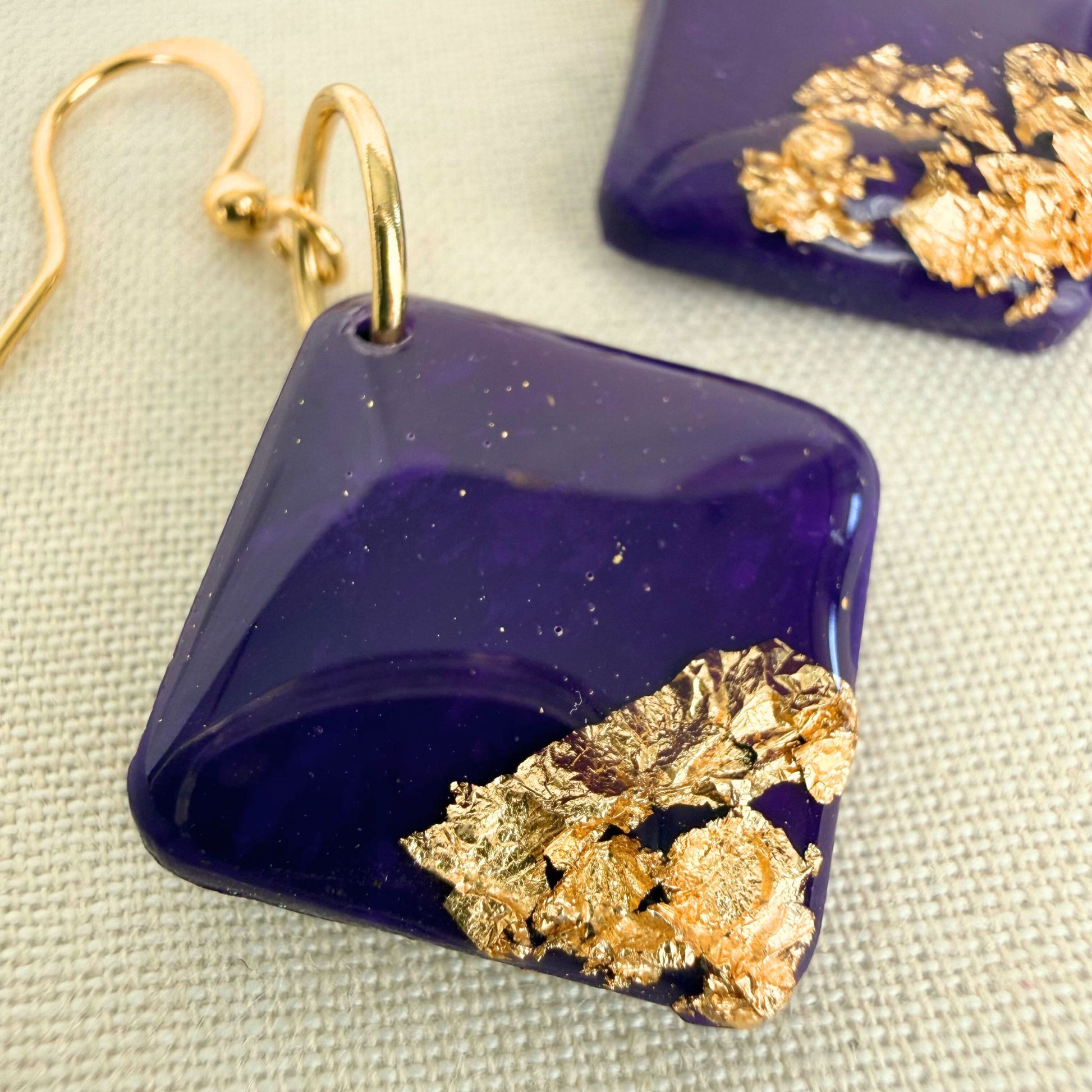 Close up of handmade dangle earrings with violet and gold design on a beige background.