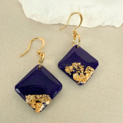 Deep violet square shaped dangle earrings with gold leaf details and gloss finish against a neutral background.