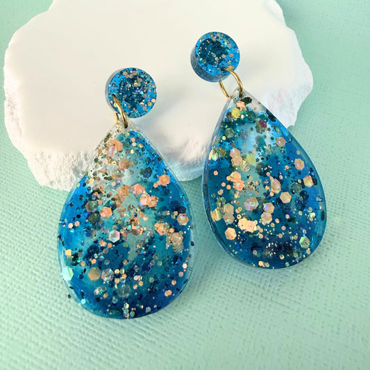 Australian handmade blue resin teardrop earrings with gold glitter accents on a light green background.