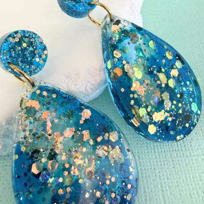 Blue resin glittery teardrop shaped drop earrings on a light blue background.