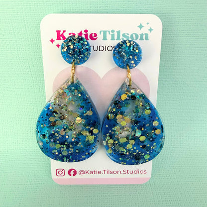 Blue teardrop drop style earrings with glitter on a Katie Tilson Studios card against a light green background.