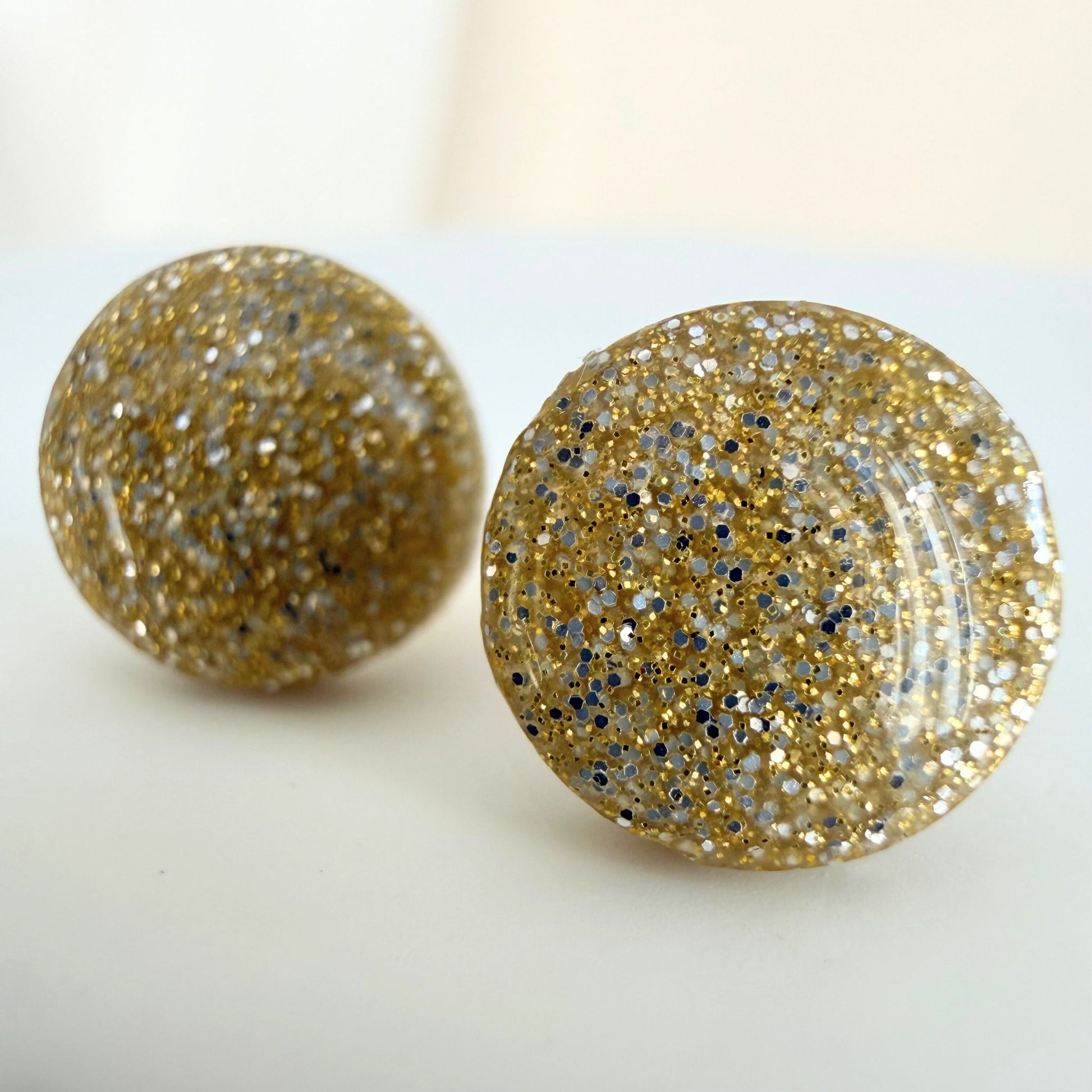 A pair of champagne gold colour circle studs with gold and silver shimmer and glossy resin finish resting on a white surface.