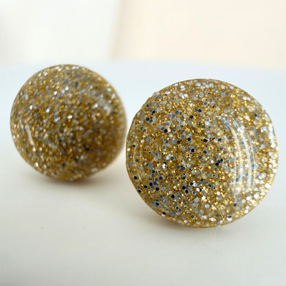 A pair of champagne gold colour circle studs with gold and silver shimmer and glossy resin finish resting on a white surface.