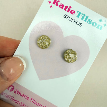 Gold glittery round stud earrings on a Katie Tilson Studios card held by a hand.