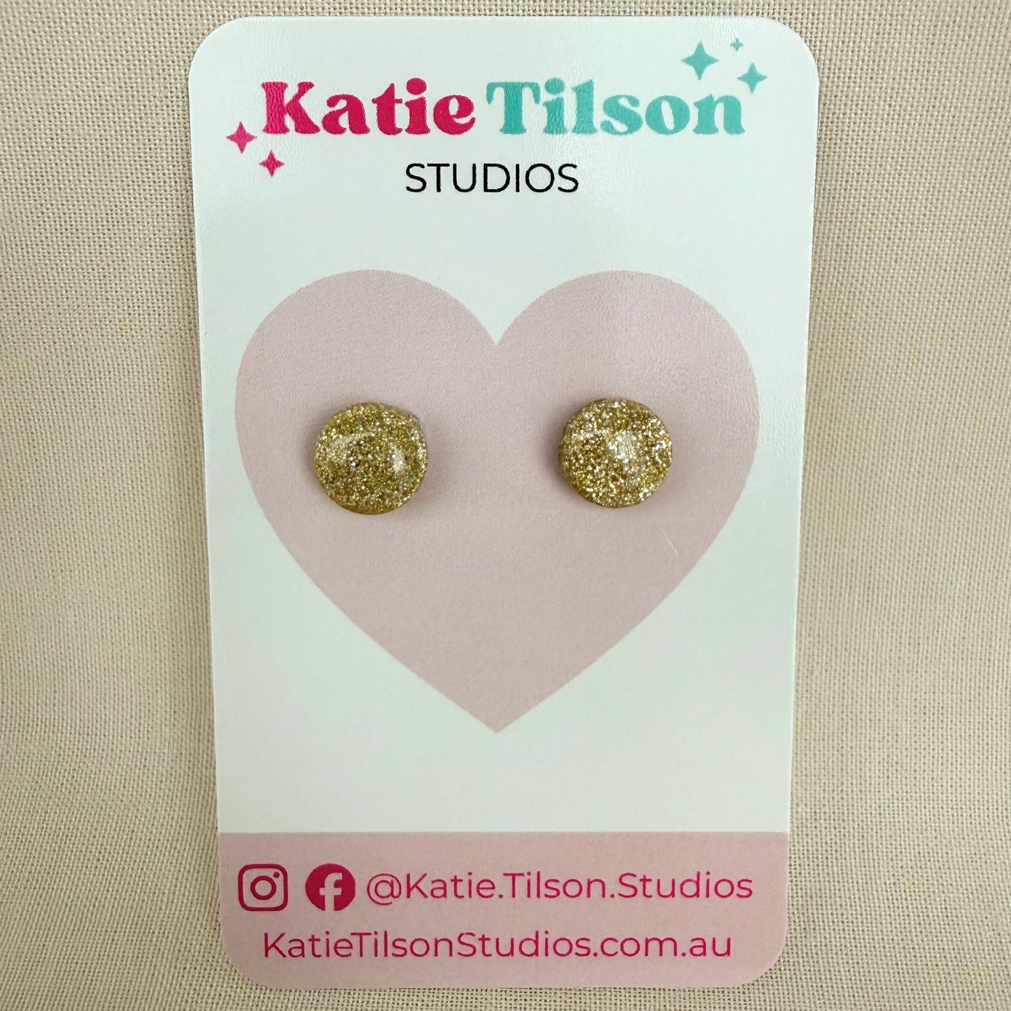 Gold glittery round stud earrings on a Katie Tilson Studios card against a beige background.