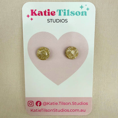 Gold glittery round stud earrings on a Katie Tilson Studios card against a beige background.