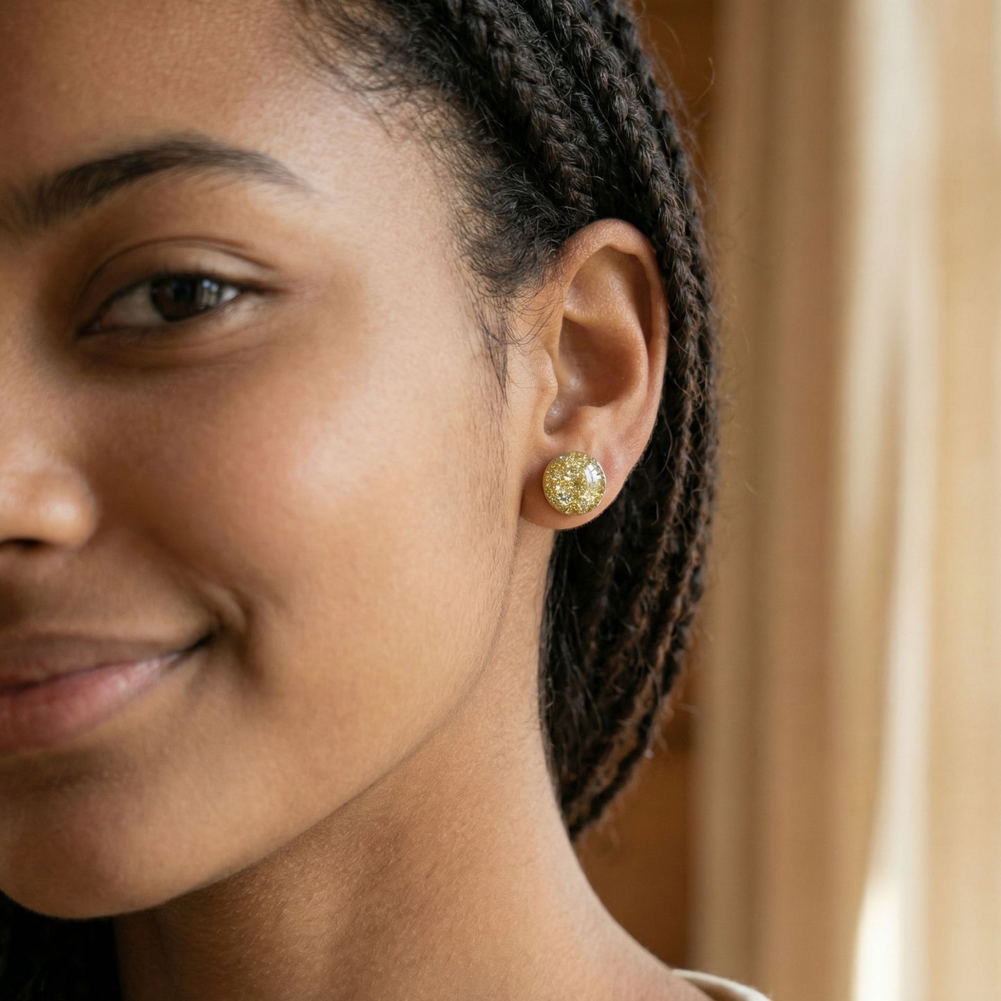 Close-up of a woman wearing round champagne gold stud earrings with a glossy finish against a blurred background.