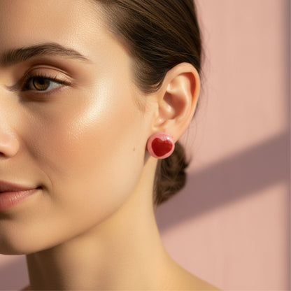 Close-up of a woman wearing red and pink stud earrings with heart detail against a pink background.