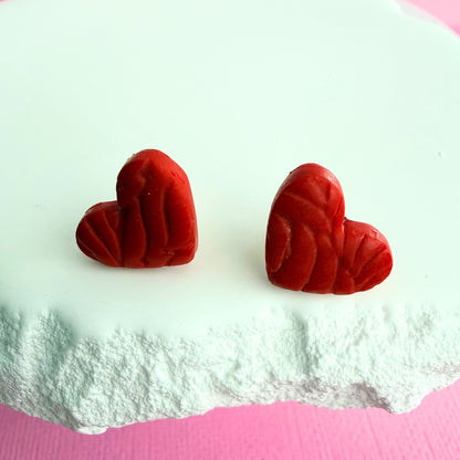 Red heart shaped stud earrings with texture on a white surface.