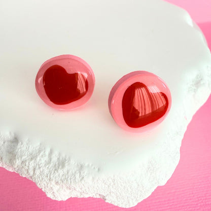 Pink handmade stud earrings with red heart details on a white stone with a pink background.