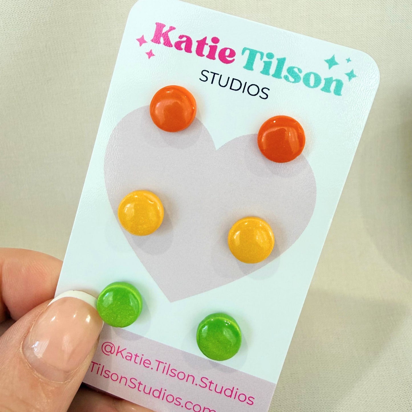 Circular stud earrings in orange, yellow and lime green displayed on a Katie Tilson Studios branded card held by a hand.