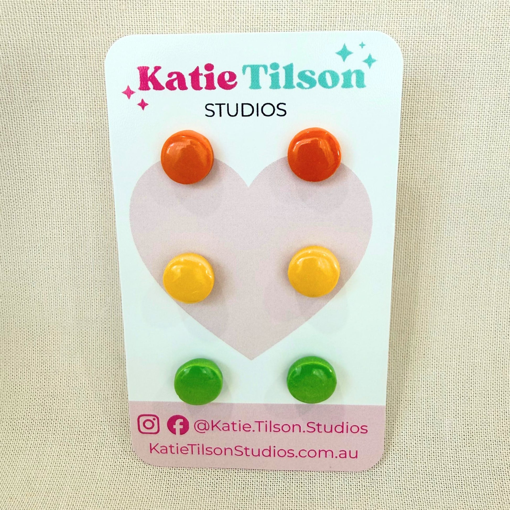 Circular stud earrings in orange, yellow and lime green displayed on a Katie Tilson Studios branded card against a beige background.